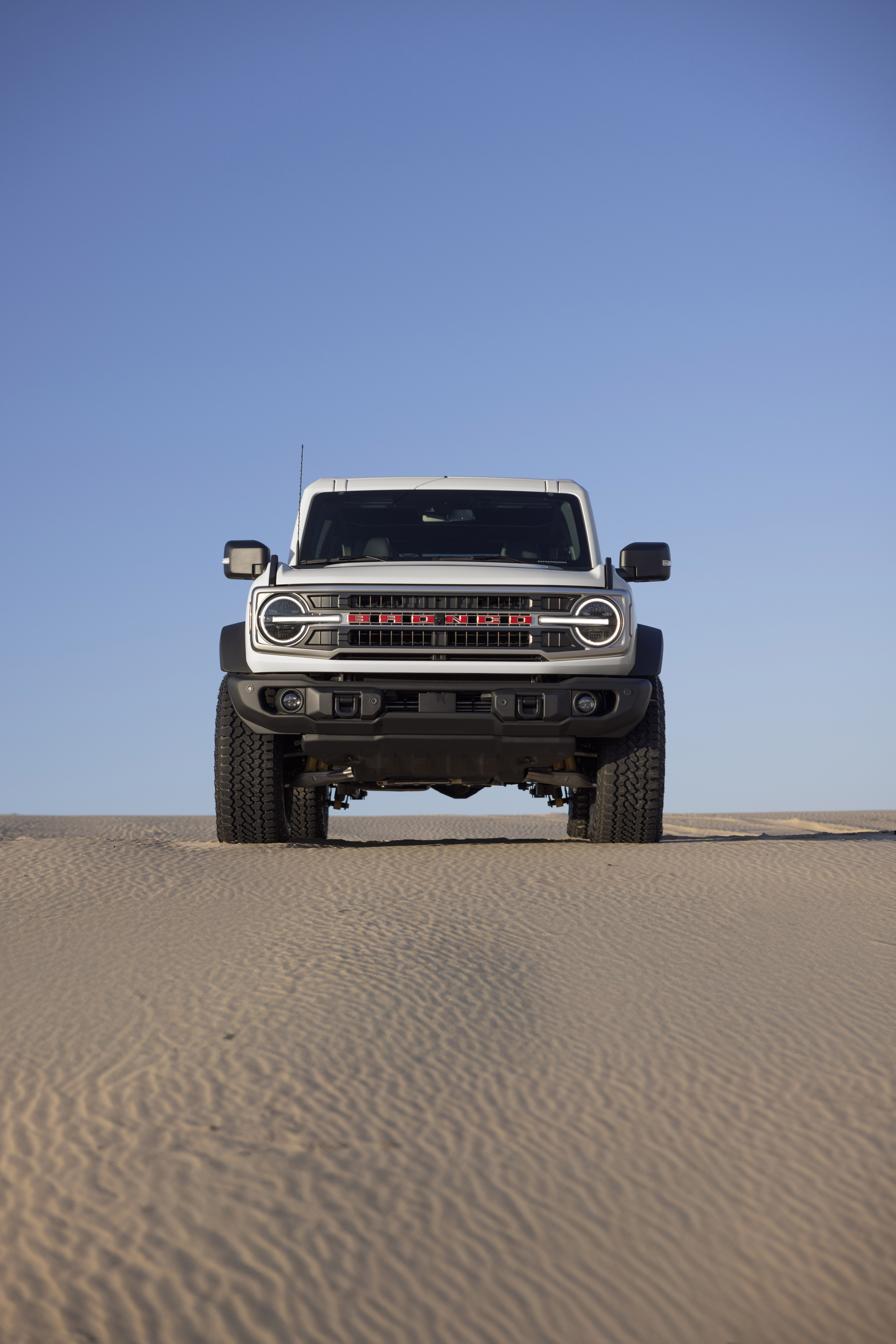 2026 Ford Bronco 60th Anniversary Package front view showing a white vehicle with "BRONCO" across the front in red letters. Vehicle is on the sand with a blue sky overhead.